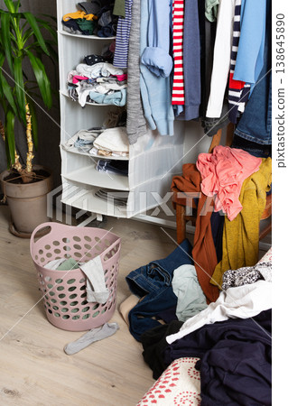 Clothes are scattered on the floor and inside a basket in a room. A chair holds additional clothes. The scene shows items ready for washing after daily use and activities 138645890