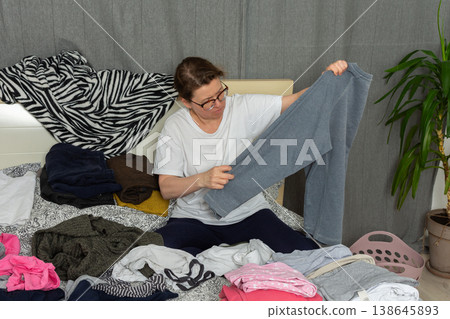 A 52-year-old woman is sorting clean laundry on a bed. She holds a pair of pants in one hand while looking at the clothes around her. A plant is seen in the corner of the room A 52-year-old woman is sorting clean laundry on a bed. She holds a pair of pants in one hand while looking at the clothes around her. A plant is seen in the corner of the room 138645893