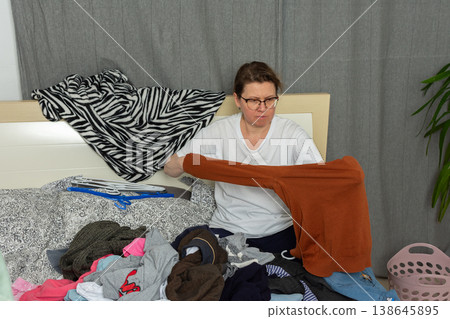 A woman, age 52, sits on her bed in a bedroom while sorting through clean laundry. She holds a piece of clothing in her hands as she organizes her belongings on the bed 138645895