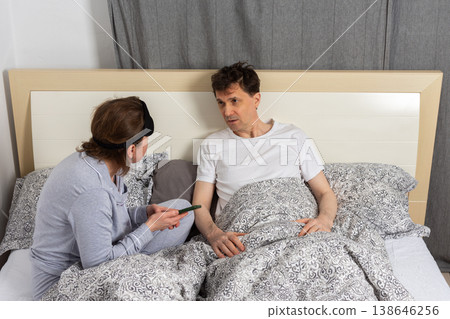A married couple, both in their fifties, wakes up in bed. The man sits up while the woman looks at her phone, sharing a moment together in their bedroom A married couple, both in their fifties, wakes up in bed. The man sits up while the woman looks at her phone, sharing a moment together in their bedroom 138646256