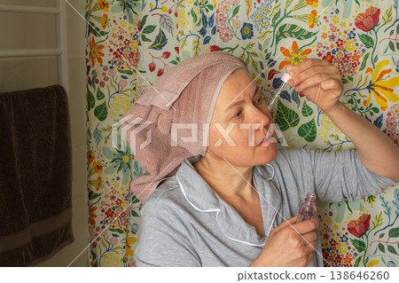 A woman in her fifties is in the bathroom applying facial serum. She wears a towel on her head and soft pajamas. It is morning, and colorful patterns surround her A woman in her fifties is in the bathroom applying facial serum. She wears a towel on her head and soft pajamas. It is morning, and colorful patterns surround her 138646260