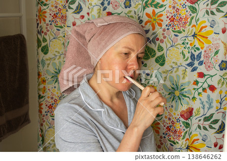A 50-year-old woman brushes her teeth in a bright bathroom. She wears a towel on her head and sits in front of a floral background. It is morning and she is starting her day 138646262
