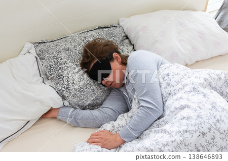 A 50-year-old woman is peacefully sleeping in a cozy bed. She wears a sleep mask and is snuggled under soft blankets, creating a serene atmosphere for rest A 50-year-old woman is peacefully sleeping in a cozy bed. She wears a sleep mask and is snuggled under soft blankets, creating a serene atmosphere for rest 138646893