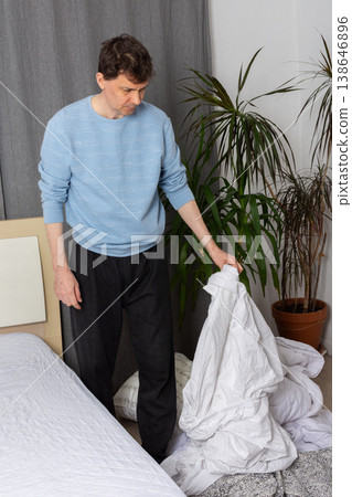 A 53-year-old man is changing the bed linen in his bedroom. He is lifting the duvet and arranging the bed for the next night. The room has a simple design and some plants 138646896