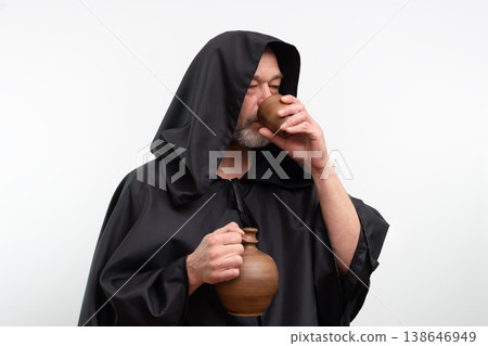 Bearded mature monk with hood drinking beverage from ceramic cup in studio. Bearded mature monk with hood drinking beverage from ceramic cup in studio. 138646949