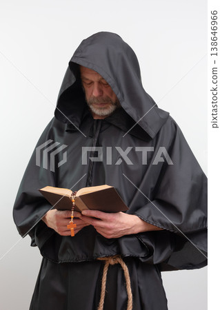 A bearded monk in a hood reads a text while holding a rosary against a white background. 138646966
