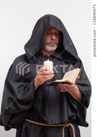 Bearded monk in hood reading text by candle light. 138646973