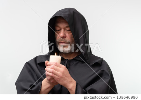 Old male monk in black hood holding candle isolated on white background. 138646980