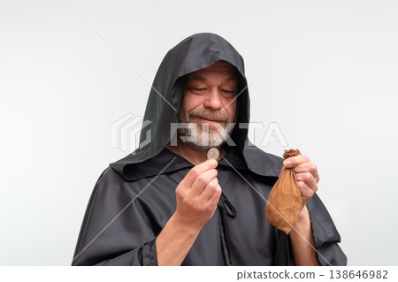 An elderly bearded monk holds a coin and a bag of money against a white background. 138646982