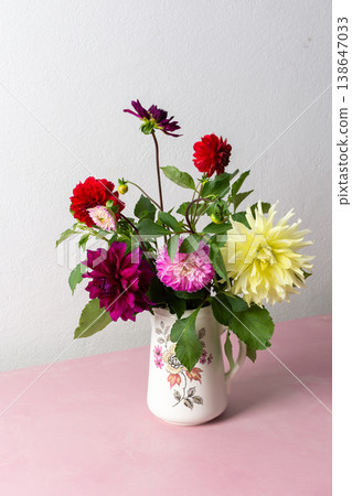 A vase holds a variety of fresh flowers with different colors. The flowers are placed on a pink table. Behind the vase is a plain wall in a bright room A vase holds a variety of fresh flowers with different colors. The flowers are placed on a pink table. Behind the vase is a plain wall in a bright room 138647033