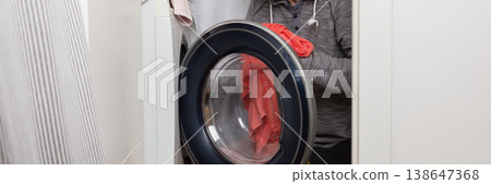 A 53-year-old man stands next to a washing machine. He is picking up clothes from the washer and placing them into a laundry basket in a home setting, banner A 53-year-old man stands next to a washing machine. He is picking up clothes from the washer and placing them into a laundry basket in a home setting, banner 138647368