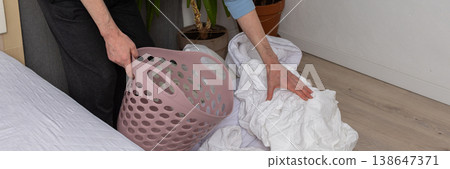 A 53-year-old man is in his bedroom folding and changing bed linen. He is holding a laundry basket while arranging the sheets on the bed. Plants are visible in the room, banner A 53-year-old man is in his bedroom folding and changing bed linen. He is holding a laundry basket while arranging the sheets on the bed. Plants are visible in the room, banner 138647371