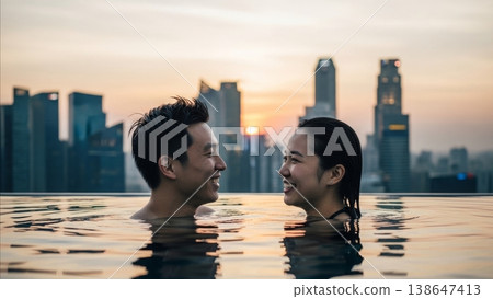 Happy Asian couple laughing together in infinity pool at sunset with modern city skyline background during romantic vacation 138647413