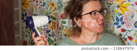 A woman prepares for the day in her bathroom. She is using a hair dryer to style her hair while standing in front of a vibrant shower curtain. Morning light fills the room, banner A woman prepares for the day in her bathroom. She is using a hair dryer to style her hair while standing in front of a vibrant shower curtain. Morning light fills the room, banner 138647974