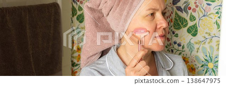 A 50 year old woman applies a facial roller to her face while standing in a bright bathroom. She has a towel wrapped around her hair and wears comfortable clothes, banner 138647975