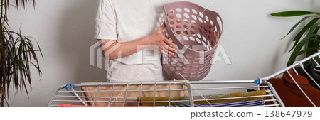 A 50-year-old woman hangs freshly washed laundry on a drying rack inside her room. She is holding a laundry basket filled with clothes, organizing her space, banner A 50-year-old woman hangs freshly washed laundry on a drying rack inside her room. She is holding a laundry basket filled with clothes, organizing her space, banner 138647979