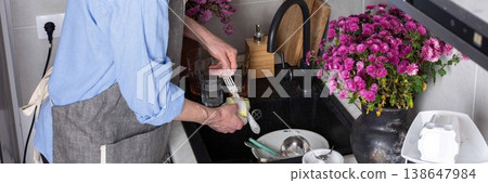 A man stands at a sink in a kitchen, washing dirty dishes and glassware. Flowers sit on the counter as fresh light comes in through the window. The setting is busy and practical, banner 138647984