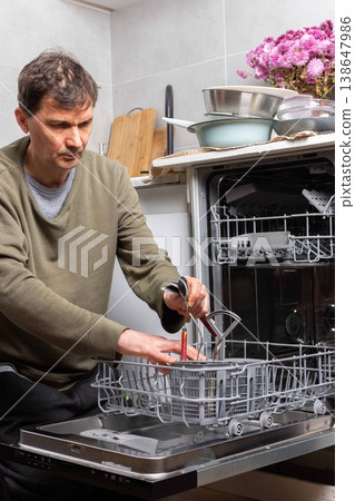 A man stands in his kitchen taking clean dishes out of the dishwasher. He focuses on the process of unloading the clean dishes and organizing them for use A man stands in his kitchen taking clean dishes out of the dishwasher. He focuses on the process of unloading the clean dishes and organizing them for use 138647986