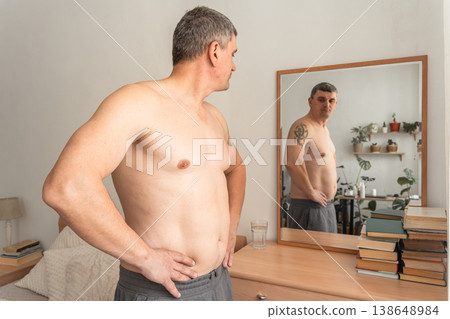 Mature man standing shirtless, looking at his reflection in a mirror, examining his changing body shape and waistline. Real person, self-acceptance, authentic lifestyle Mature man standing shirtless, looking at his reflection in a mirror, examining his changing body shape and waistline. Real person, self-acceptance, authentic lifestyle 138648984