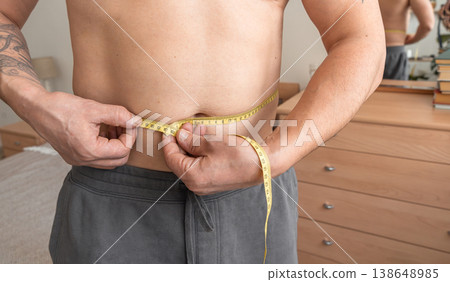 Man measuring his waist with a tape measure, checking progress on his weight loss journey in the bedroom. Real person, self-acceptance, authentic lifestyle Man measuring his waist with a tape measure, checking progress on his weight loss journey in the bedroom. Real person, self-acceptance, authentic lifestyle 138648985