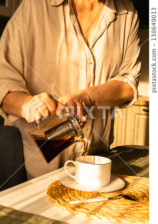 Womans hands pouring fresh brewed tea from glass teapot into cup. Warm morning light creating cozy atmosphere during breakfast. Authenticity, everyday moment Womans hands pouring fresh brewed tea from glass teapot into cup. Warm morning light creating cozy atmosphere during breakfast. Authenticity, everyday moment 138649013