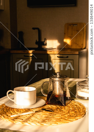 Cozy morning tea ritual: glass teapot, cup and sugar bowl on a sunlit kitchen table, warm golden-hour light and calm domestic charm. Tea time table setup for a cozy tea party at home 138649014