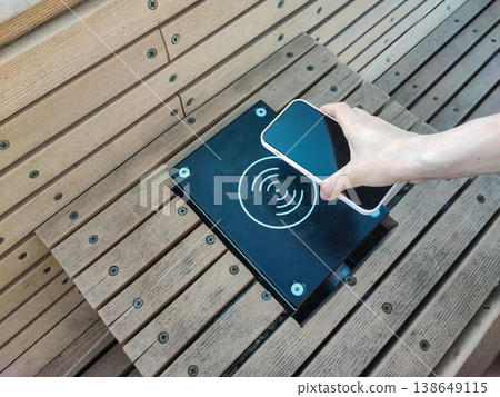 Hand holds smartphone above wireless charging pad on wooden bench outdoors during daylight 138649115