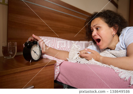 Shocked Woman Reaching Alarm Clock in Bed After Oversleeping 138649586