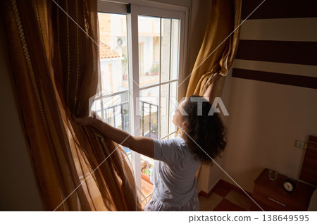 A woman standing by a window, gently opening curtains to let in the morning sunlight. A woman standing by a window, gently opening curtains to let in the morning sunlight. 138649595