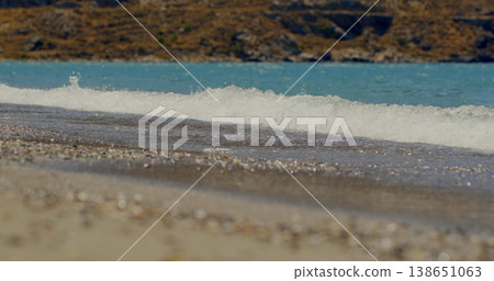 Waves crash on the shore at a beach during the midday sun with clear water and pebbles scattered on the sand 138651063