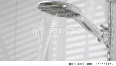 Water flows from a showerhead in a bathroom during the day, with light shining through blinds onto the wall 138651169
