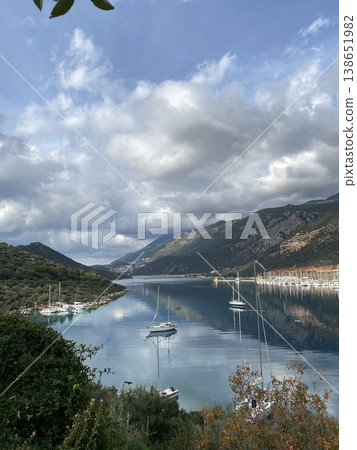 Scenic view of sailboats anchored in a calm bay surrounded by mountains and reflected clouds Scenic view of sailboats anchored in a calm bay surrounded by mountains and reflected clouds 138651982
