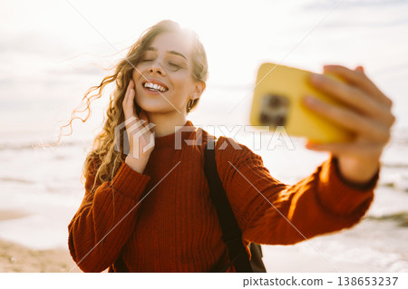 Young woman joyfully taking selfie on beach during golden hour. Selfie time. Cold windy weather. Young woman joyfully taking selfie on beach during golden hour. Selfie time. Cold windy weather. 138653237