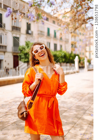 Beautiful young woman in an orange dress enjoys a sunny day in a lively setting with blooming trees Beautiful young woman in an orange dress enjoys a sunny day in a lively setting with blooming trees 138653798