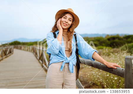 A happy young woman stands on a wooden boardwalk near the beach, wearing a straw hat and blue shirt 138653955
