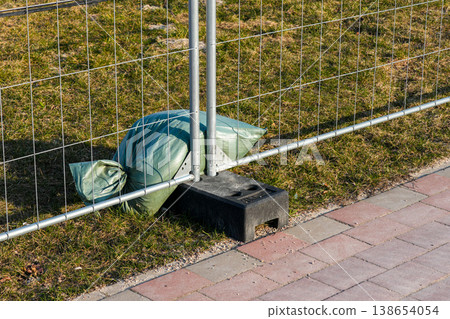Temporary construction fence secured with sandbag weight on sidewalk near grassy roadside area Temporary construction fence secured with sandbag weight on sidewalk near grassy roadside area 138654054