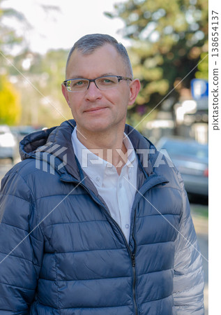 Man aged 51 wearing glasses stands outdoors with road and cars in city Man aged 51 wearing glasses stands outdoors with road and cars in city 138654137