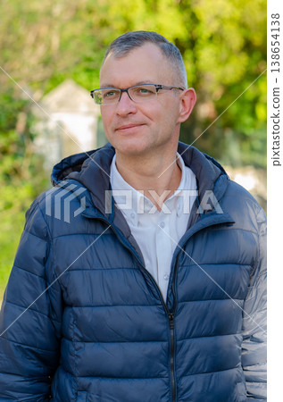 Man wearing glasses stands outdoors against greenery in jacket and looks confident Man wearing glasses stands outdoors against greenery in jacket and looks confident 138654138