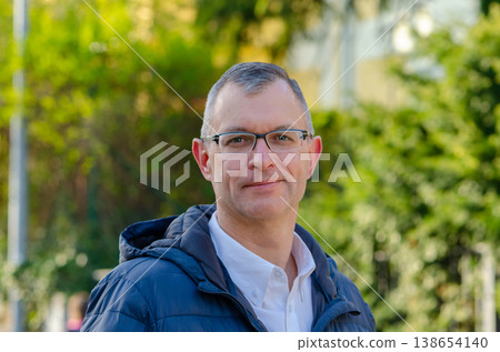 Man wearing glasses stands outdoors with green plants background in jacket and looks calm Man wearing glasses stands outdoors with green plants background in jacket and looks calm 138654140