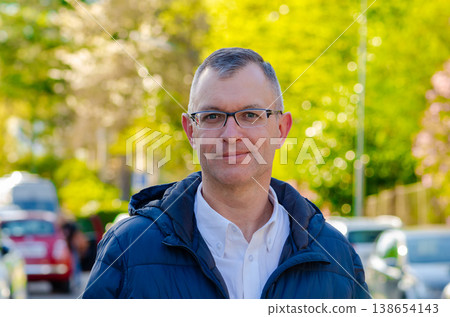 Business man wearing glasses stands outdoors in jacket with greenery and city movement Business man wearing glasses stands outdoors in jacket with greenery and city movement 138654143