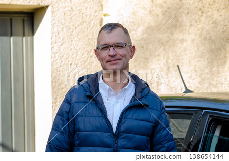 Man wearing glasses stands near car by house in jacket looking calmly at camera Man wearing glasses stands near car by house in jacket looking calmly at camera 138654144