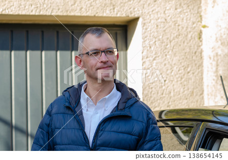 Adult man aged 51 wearing glasses poses near car outdoors with house background Adult man aged 51 wearing glasses poses near car outdoors with house background 138654145