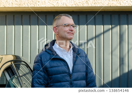 Man wearing glasses stands by garage outdoors in jacket looking into distance in daylight Man wearing glasses stands by garage outdoors in jacket looking into distance in daylight 138654146