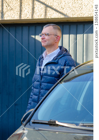 Adult man aged 51 wearing glasses poses near car with building background Adult man aged 51 wearing glasses poses near car with building background 138654148