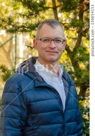 Adult man wearing glasses stands outdoors near conifer tree and looks at camera with confident expression Adult man wearing glasses stands outdoors near conifer tree and looks at camera with confident expression 138654153