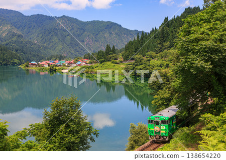 A special Tadami Line train speeds through the landscape against the backdrop of the Tadami River, reflecting the bright blue summer sky and white clouds, and the Taishi settlement with its rows of colorful roofs. A special Tadami Line train speeds through the landscape against the backdrop of the Tadami River, reflecting the bright blue summer sky and white clouds, and the Taishi settlement with its rows of colorful roofs. 138654220