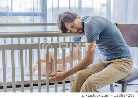 Tired father sitting by baby crib at home watching sleeping infant. Exhausted dad caring for newborn at night. Parenting fatigue, childcare responsibility, love and real family life concept 138654292