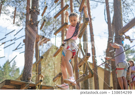 A girl with a safety rope walks along a rope, passing obstacles on a suspended bridge. A girl with a safety rope walks along a rope, passing obstacles on a suspended bridge. 138654299
