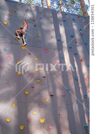 A girl with a safety rope climbs the steep wall of a climbing wall located in a pine park. Extreme sports in an amusement park. A girl with a safety rope climbs the steep wall of a climbing wall located in a pine park. Extreme sports in an amusement park. 138654301