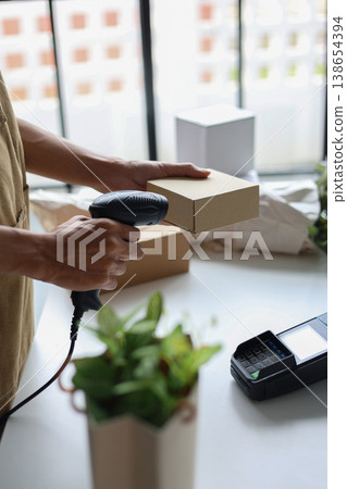 A person using a barcode scanner to scan a small cardboard box in a retail shop for inventory management or preparing an order for shipping. A person using a barcode scanner to scan a small cardboard box in a retail shop for inventory management or preparing an order for shipping. 138654394